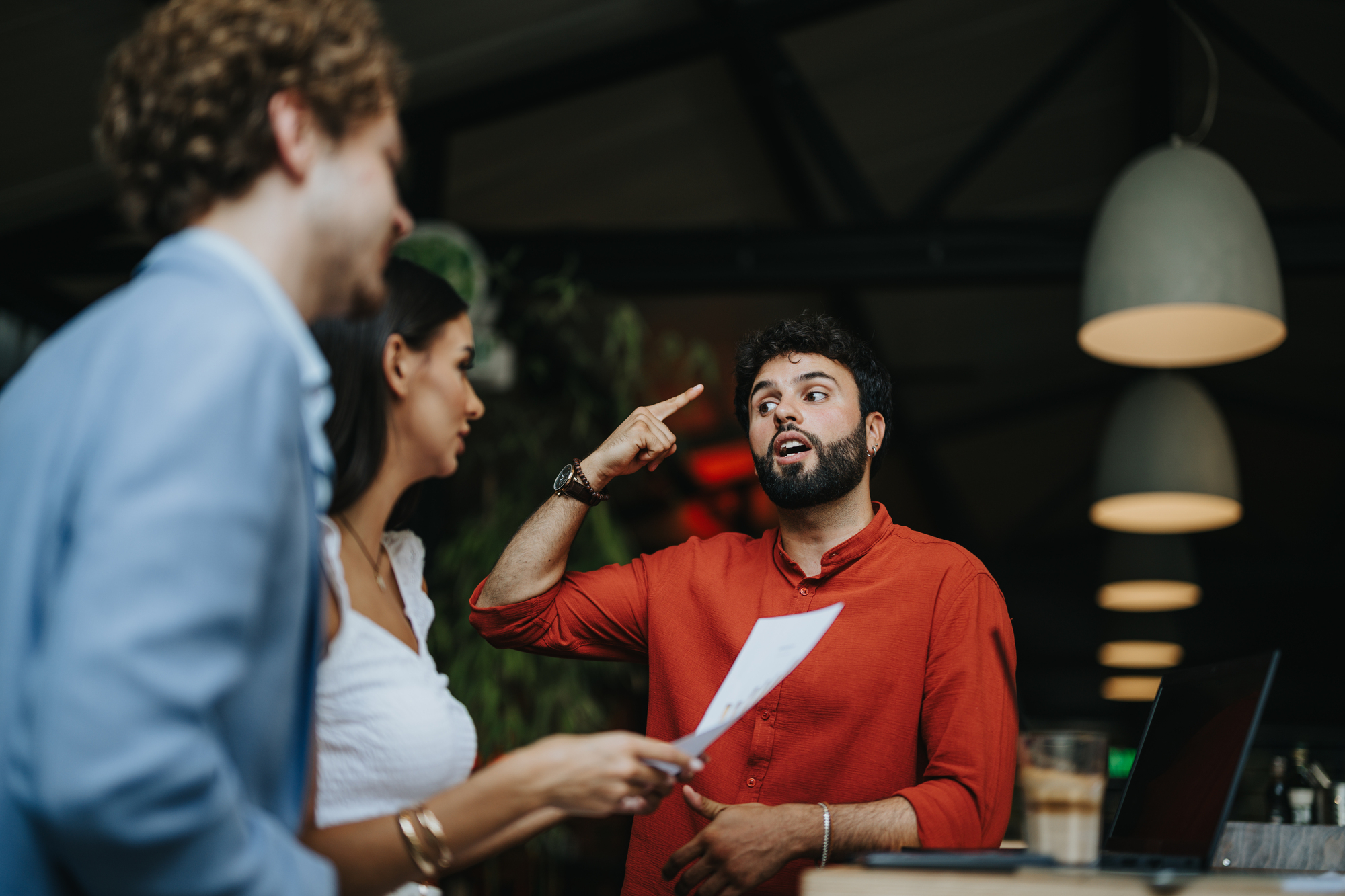 A man in a red shirt enthusiastically gestures while holding a piece of paper in a social setting. Two other people, a woman and a man, stand nearby, engaged in conversation. They are indoors, with modern pendant lights overhead.