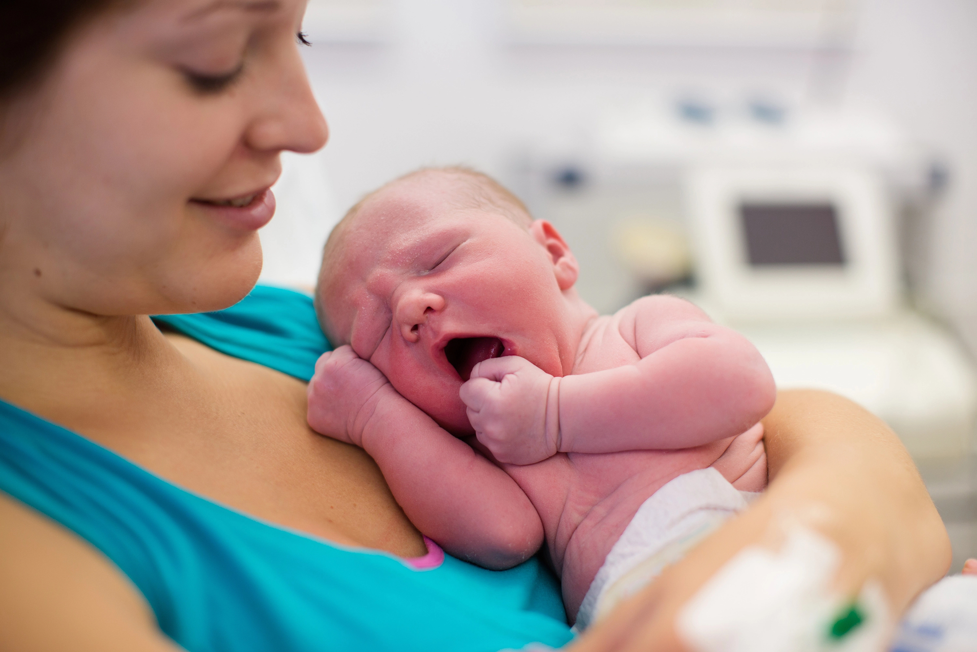 A woman smiles while holding a newborn baby who is yawning. The baby is wrapped in a diaper and nestled closely to the woman's chest in a hospital setting. The background is softly blurred.