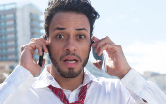 A surprised man with a beard and mustache holds two smartphones to his ears. He wears a white shirt and a red tie, standing outdoors with a blurred cityscape in the background.