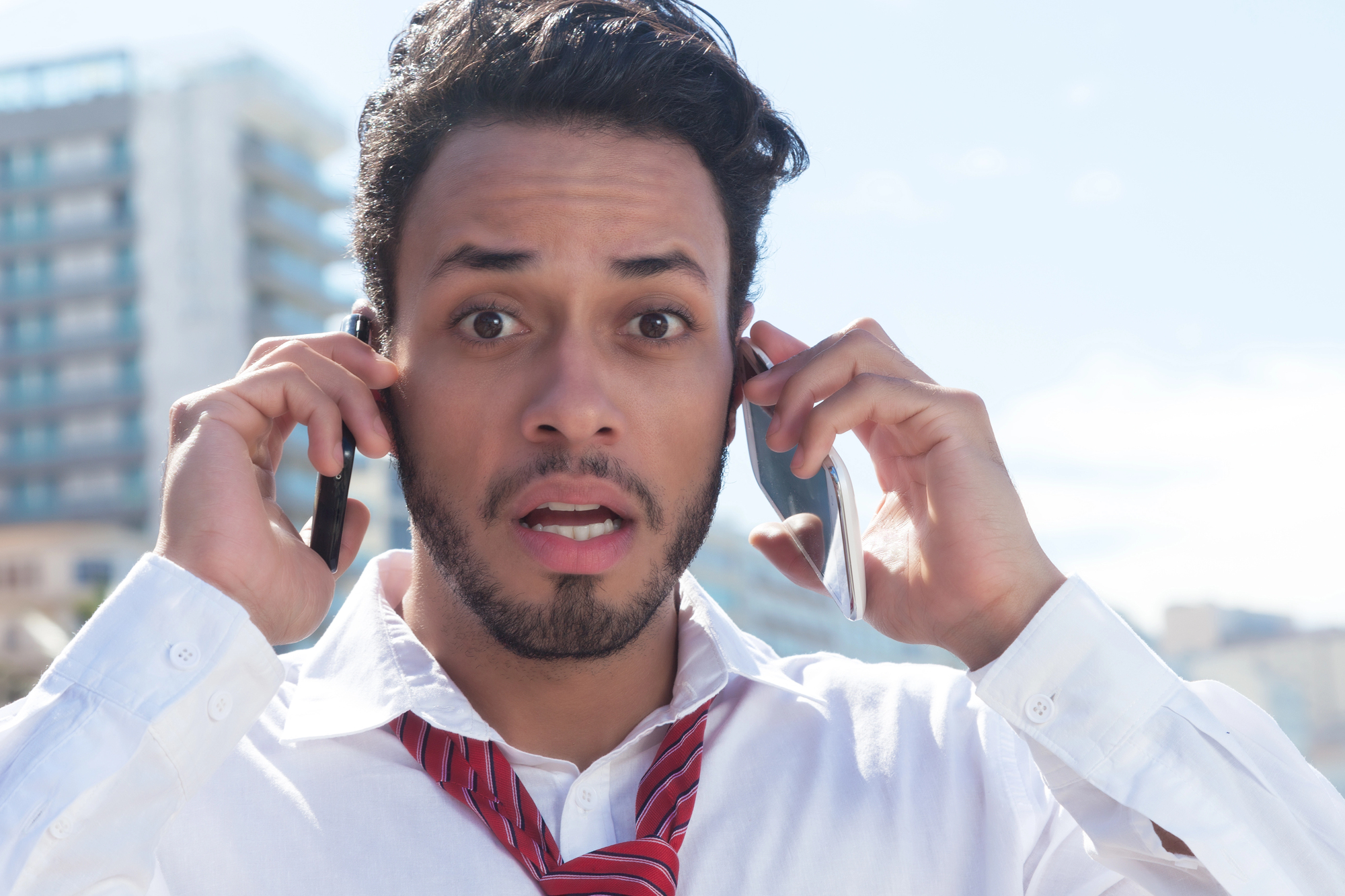 A surprised man with a beard and mustache holds two smartphones to his ears. He wears a white shirt and a red tie, standing outdoors with a blurred cityscape in the background.