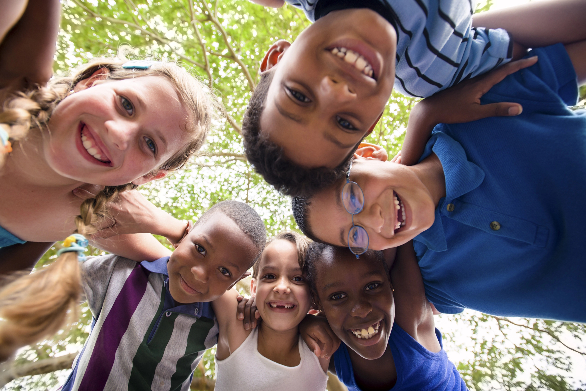 A group of smiling children huddled together, looking down at the camera under a canopy of green trees. They appear joyful and are dressed in colorful summer attire, creating a lively and cheerful atmosphere.