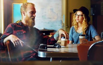 A man with a beard and a woman wearing glasses and a hat sit at a table in a cozy cafe, each holding a cup of coffee. They are engaged in a lively conversation, surrounded by warm lighting and a blurred background.