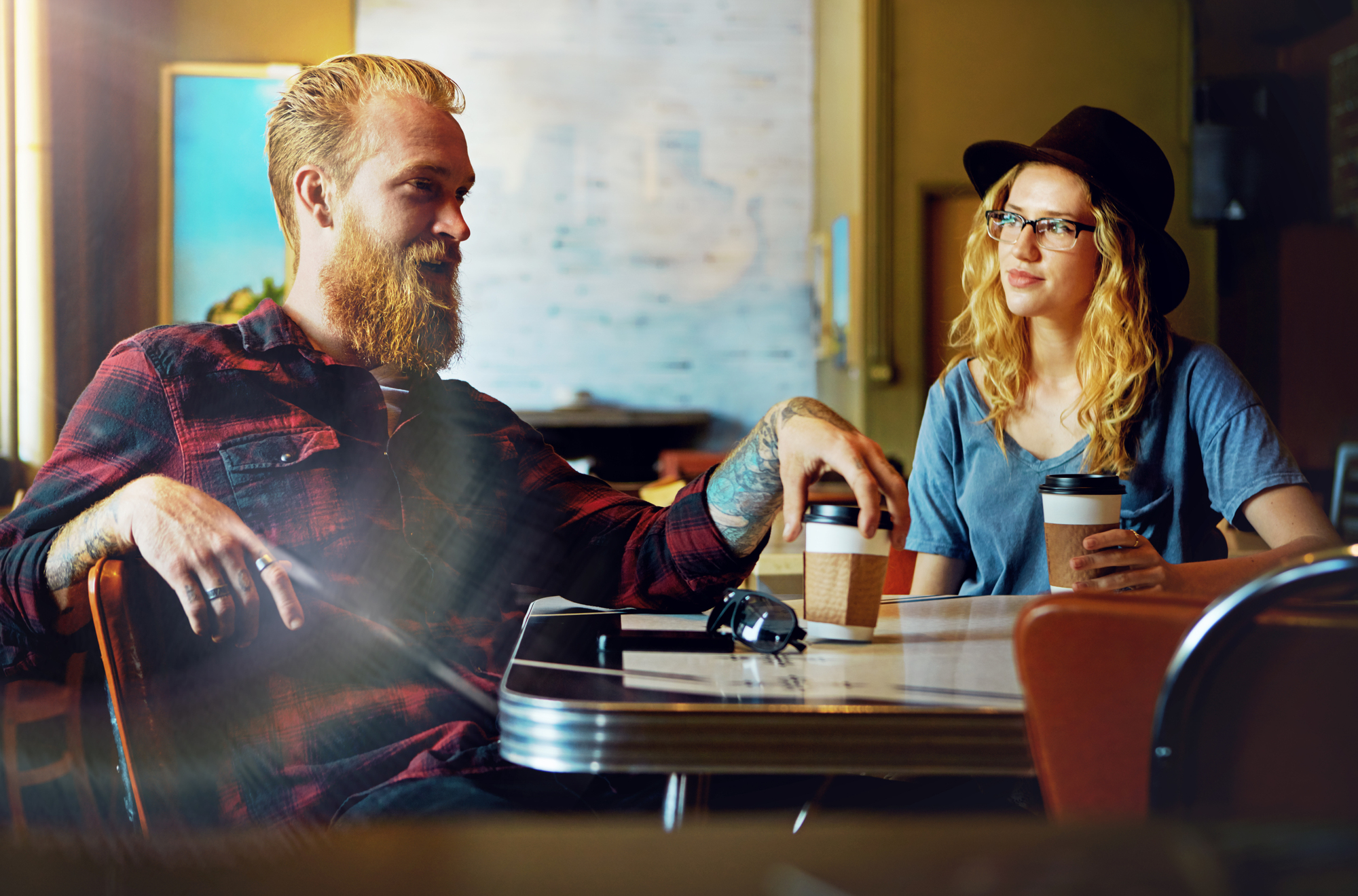 A man with a beard and a woman wearing glasses and a hat sit at a table in a cozy cafe, each holding a cup of coffee. They are engaged in a lively conversation, surrounded by warm lighting and a blurred background.