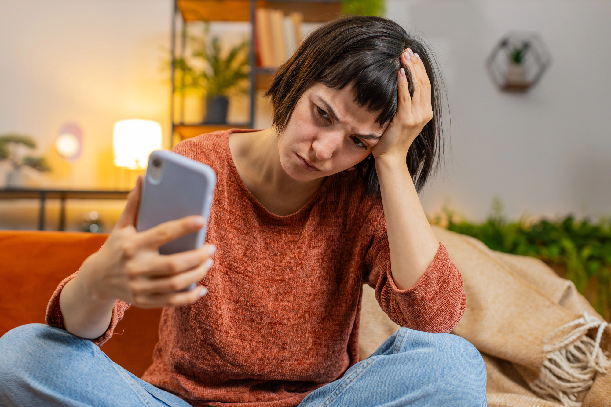 A woman with short dark hair, wearing a red sweater, sits on a couch holding a smartphone. She rests her head on one hand, looking stressed. The background shows a cozy room with shelves and plants.