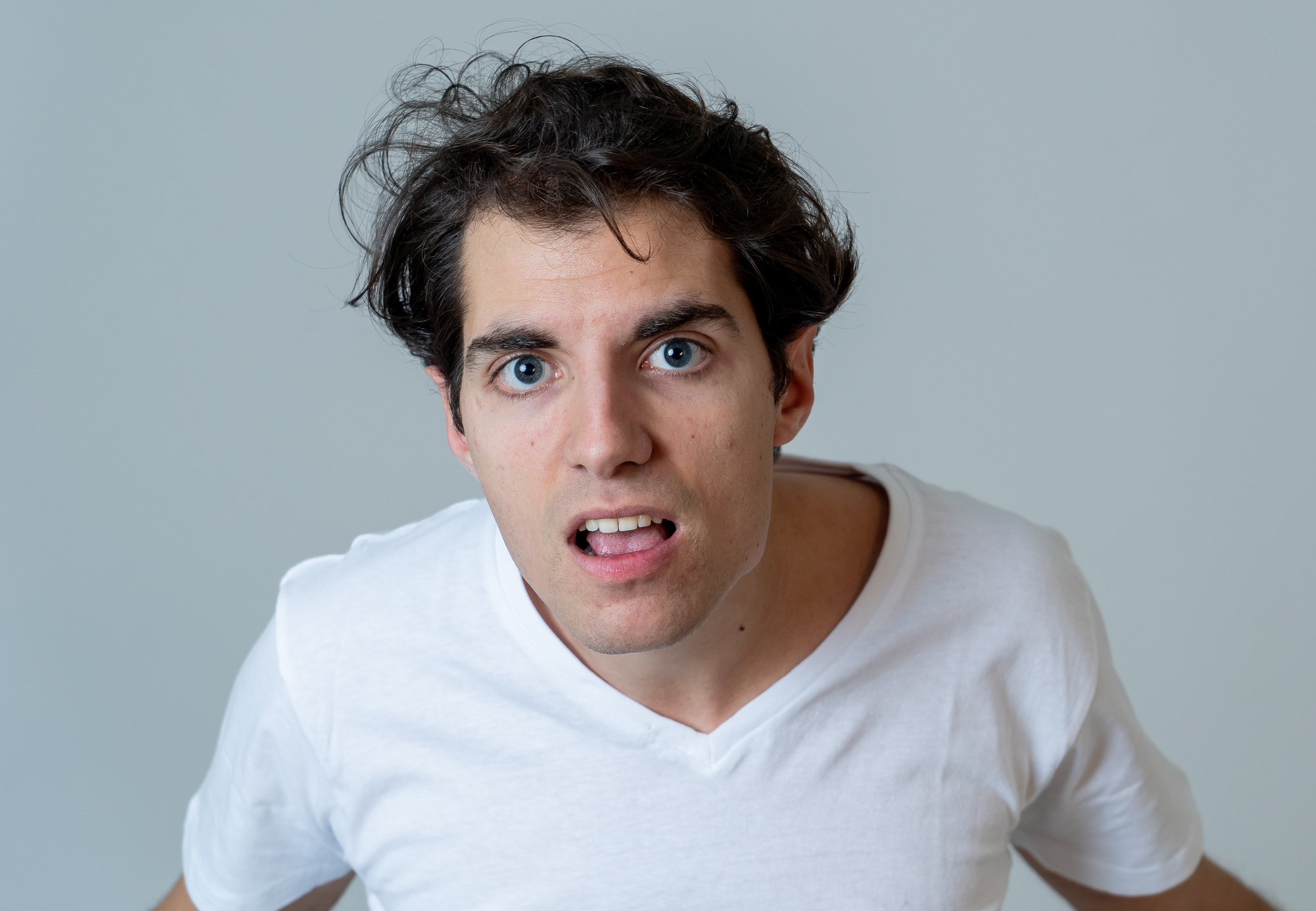 A man with messy dark hair and blue eyes looks into the camera with a confused expression, wearing a white T-shirt. The background is a plain light gray.