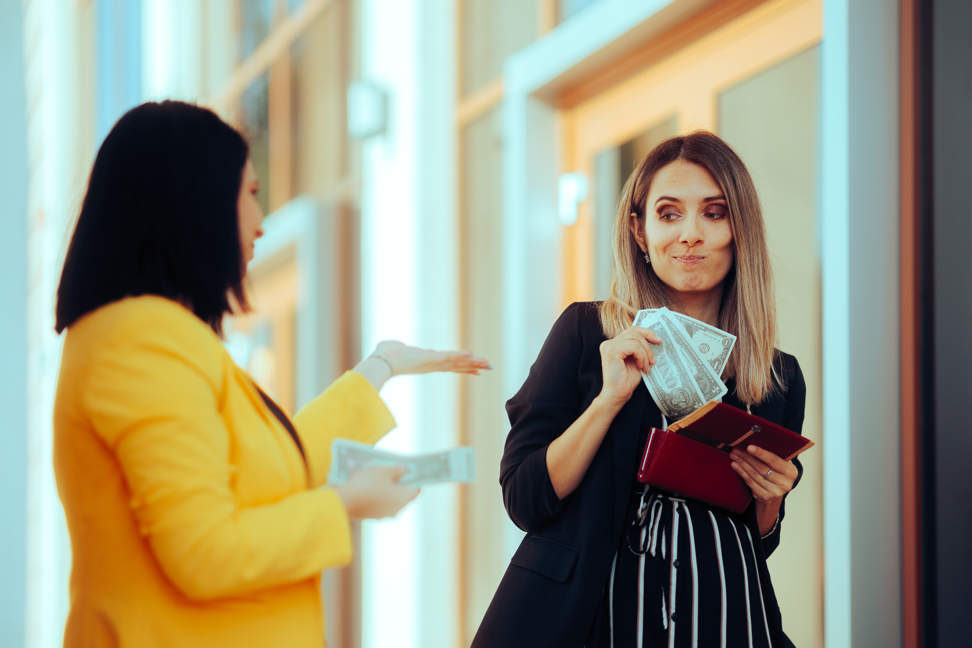 Two women are interacting. One, in a yellow blazer, gestures while showing money. The other, in a black outfit, holds dollar bills and a wallet, looking amused. They're standing outdoors near a building.