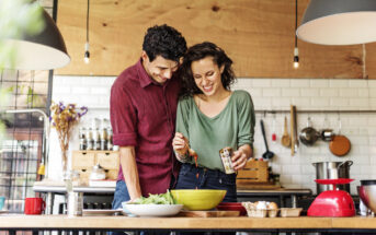 A couple cooking together in a modern kitchen. The man, in a red shirt, leans over a table with vegetables, while the woman, in a green top, stirs something in a bowl. They both smile and are surrounded by kitchen utensils and ingredients.