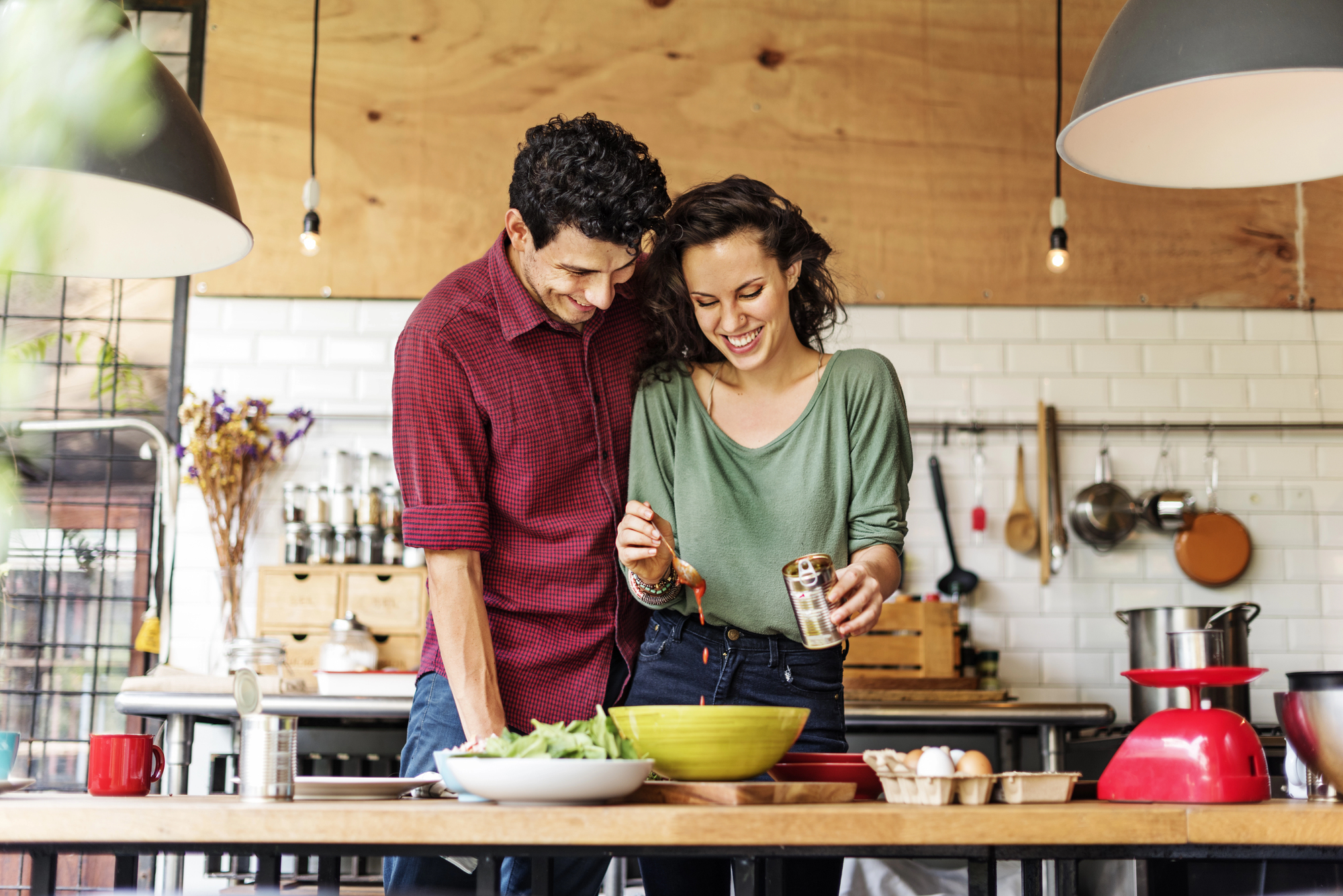 A couple cooking together in a modern kitchen. The man, in a red shirt, leans over a table with vegetables, while the woman, in a green top, stirs something in a bowl. They both smile and are surrounded by kitchen utensils and ingredients.