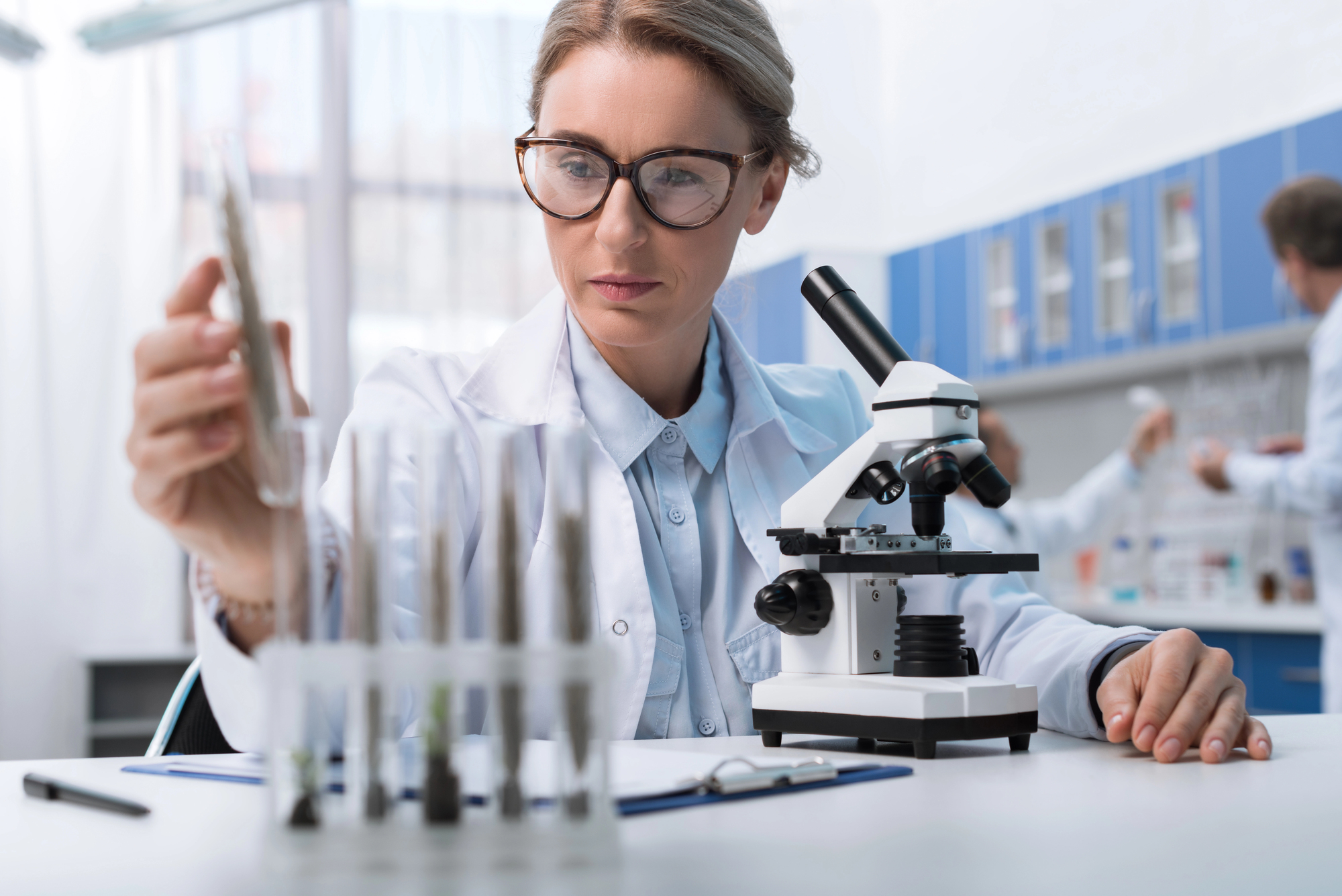 A scientist in a lab coat examines a test tube in a laboratory setting. A microscope is positioned on the table, and other lab equipment is visible in the background. Two colleagues are working at benches in the distance.