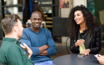 Four people sit at an outdoor café table, engaged in conversation and smiling. One person is holding a phone, while the others appear relaxed and attentive, creating a casual and friendly atmosphere.