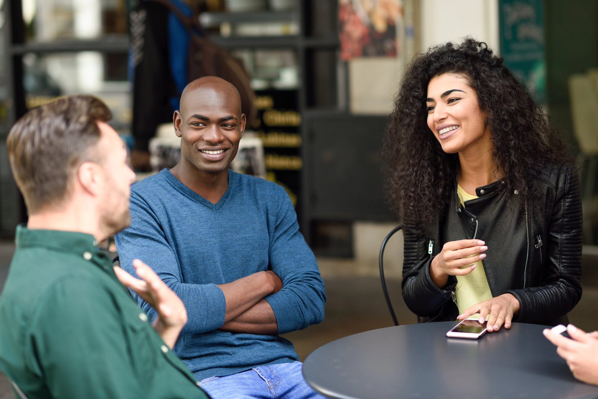 Four people sit at an outdoor café table, engaged in conversation and smiling. One person is holding a phone, while the others appear relaxed and attentive, creating a casual and friendly atmosphere.