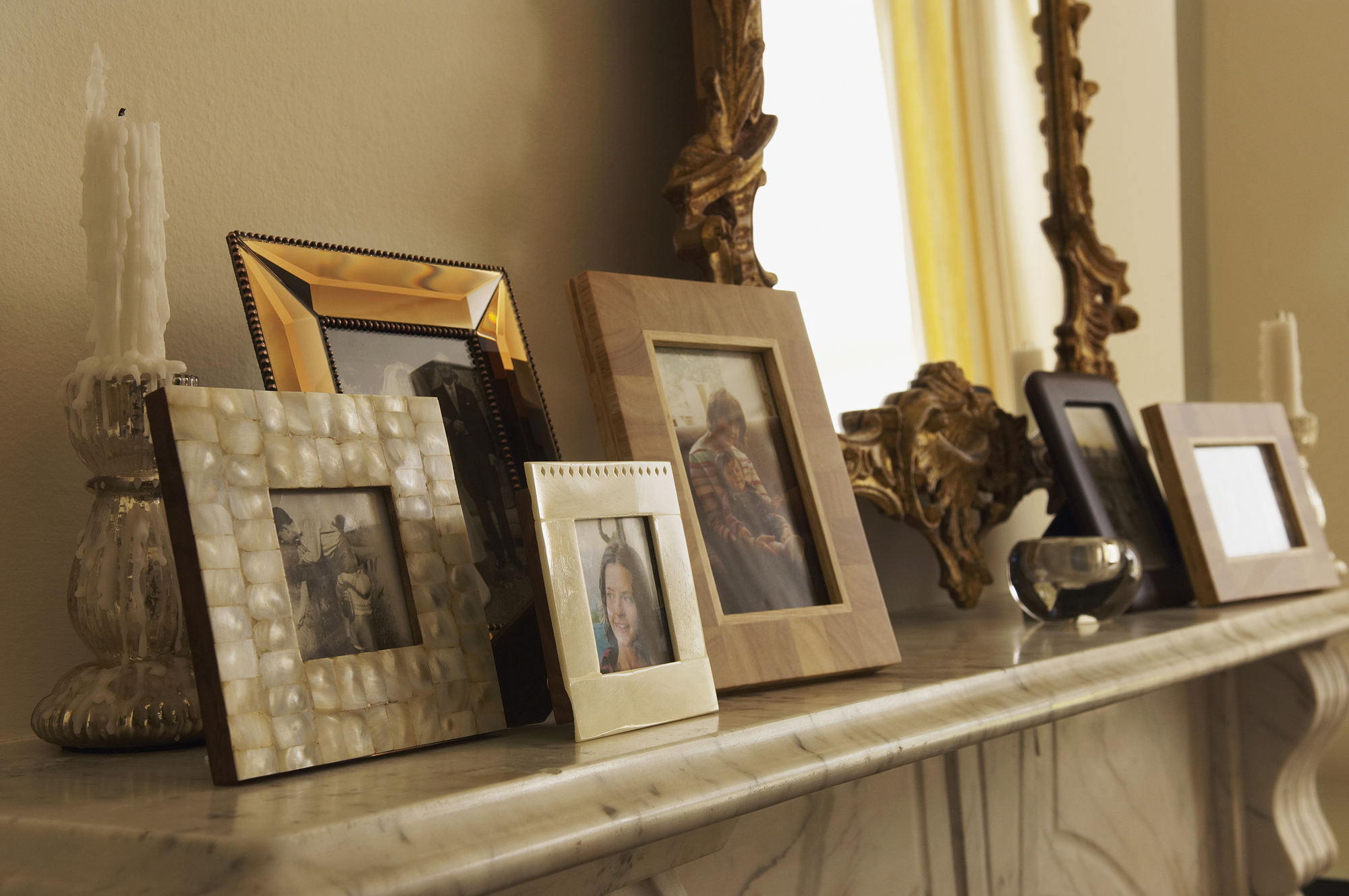 A collection of framed photographs and tall, partly melted candles are arranged on a marble mantelpiece. A large ornate mirror reflects part of the room in the background.