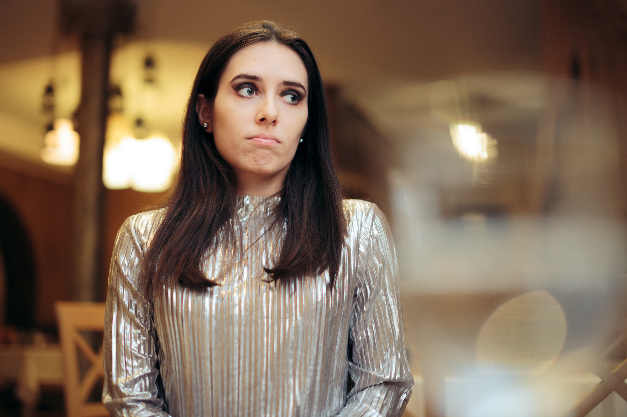 A woman with long brown hair, wearing a shiny silver top, sits indoors. She has a thoughtful expression and is looking slightly to the side. The background is softly blurred, and the scene has warm lighting.