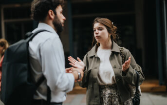 A man and a woman are engaged in conversation on a city street. The man wears a white shirt and has a backpack, while the woman has a braided hairstyle and is gesturing with her hands, wearing a light jacket. The background is slightly blurred.