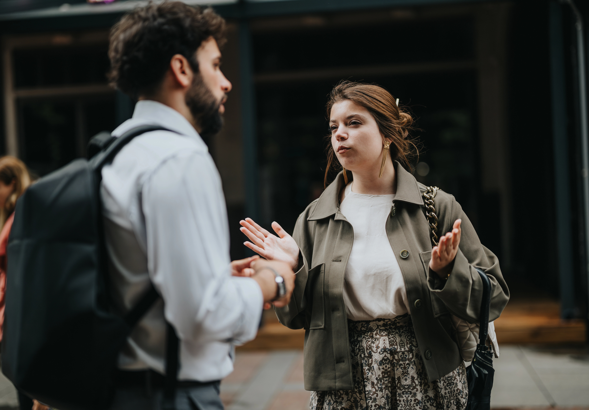 A man and a woman are engaged in conversation on a city street. The man wears a white shirt and has a backpack, while the woman has a braided hairstyle and is gesturing with her hands, wearing a light jacket. The background is slightly blurred.