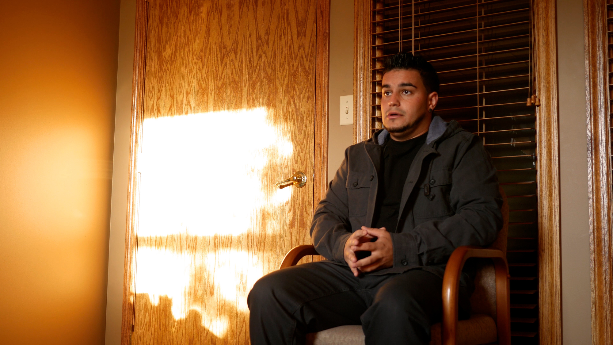 A person sits in a chair in a warmly lit room, hands clasped, looking thoughtfully forward. Sunlight streams through a window, casting bright patterns on a wooden door. The atmosphere is calm and contemplative.