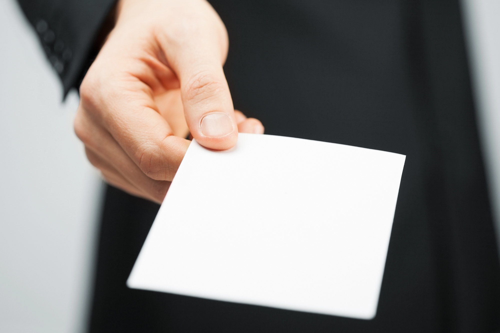 A person in a black suit extends their hand holding a blank white card towards the viewer. The background is out of focus, emphasizing the hand and card.