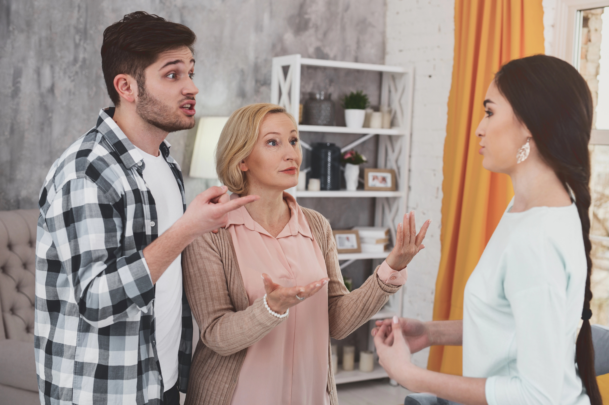 A young man and an older woman are engaged in a discussion with a young woman in a living room. The young man appears expressive, the older woman seems concerned, and the young woman is listening. Bookshelves and a curtain are in the background.