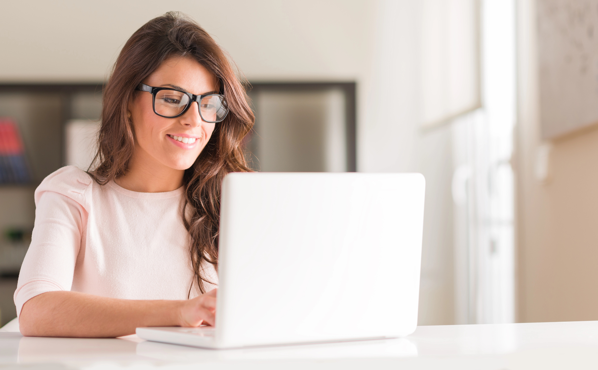 A woman with glasses is smiling while working on a laptop at a bright, modern desk. She is wearing a light pink shirt and has long brown hair. The background features blurred shelves and a large window.