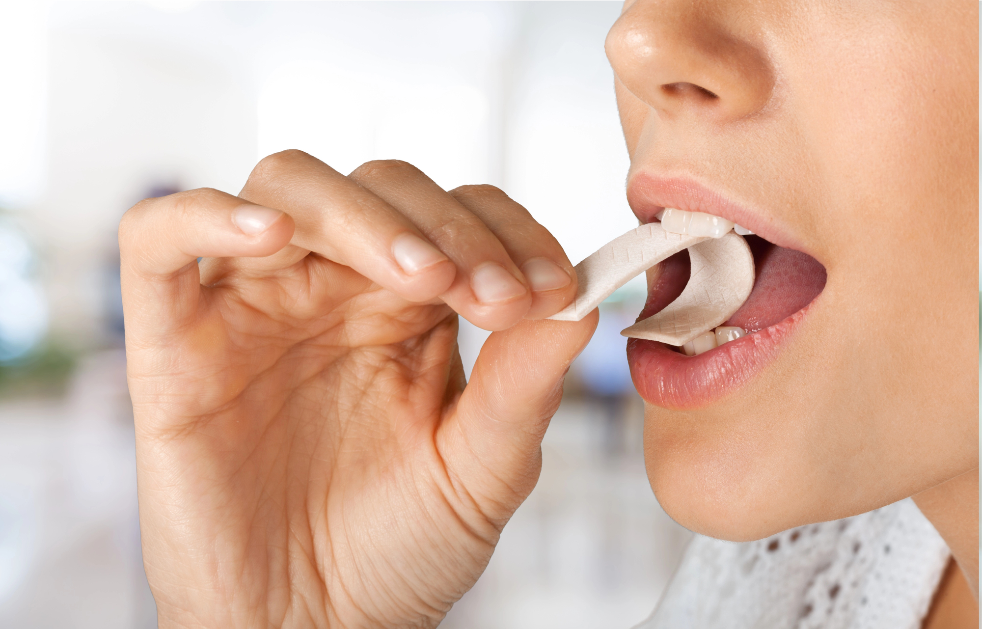 A close-up of a person placing a piece of gum in their mouth with their thumb and index finger. The background is softly blurred, emphasizing the action.