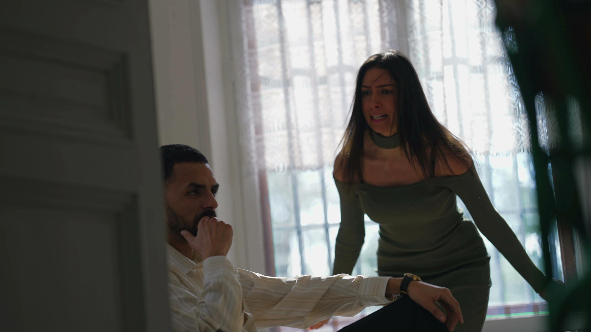 A woman in a green dress speaks intensely to a man seated nearby in a dimly lit room with sheer curtains. The man appears thoughtful, resting his chin on his hand, looking away from her.