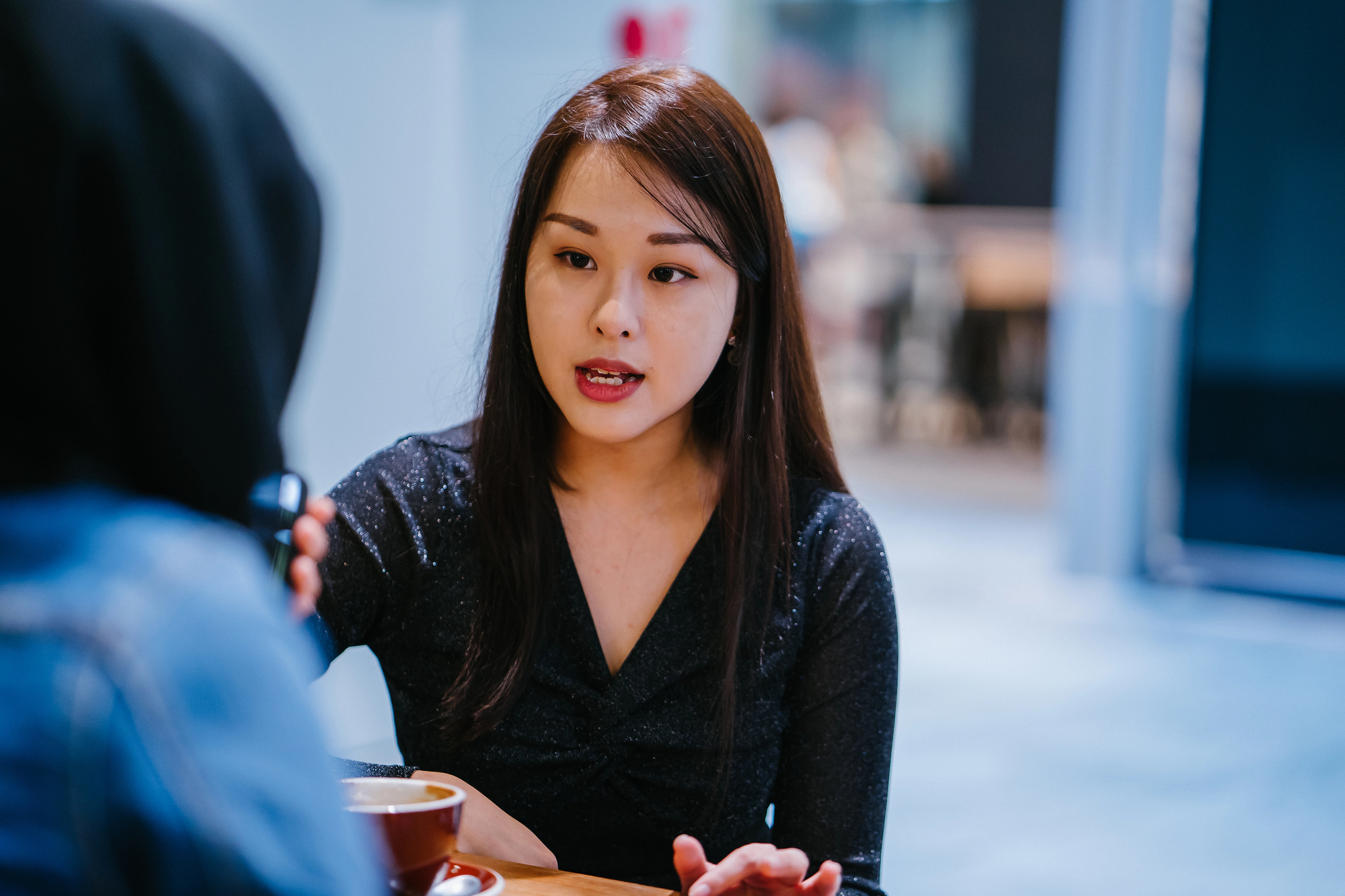 A woman with long hair, wearing a black top, is engaged in a conversation with another person across a table. A cup is in front of her. The background is a blurred indoor setting.