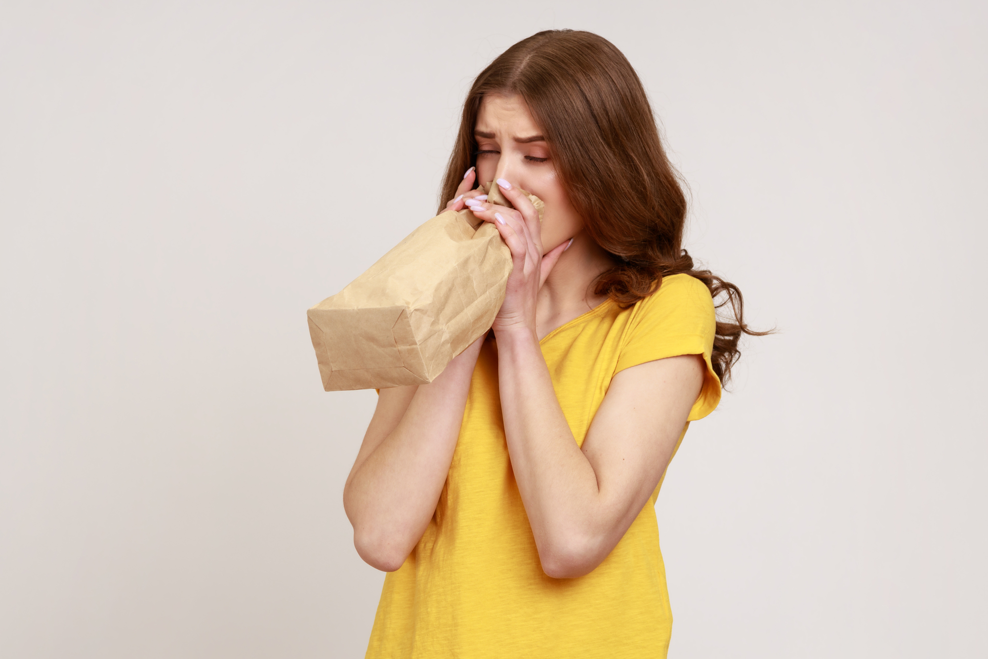 A woman with long brown hair wearing a yellow shirt is standing against a plain background. She is holding a paper bag close to her mouth, appearing to be breathing into it. Her eyes are slightly closed, conveying a sense of anxiety or distress.