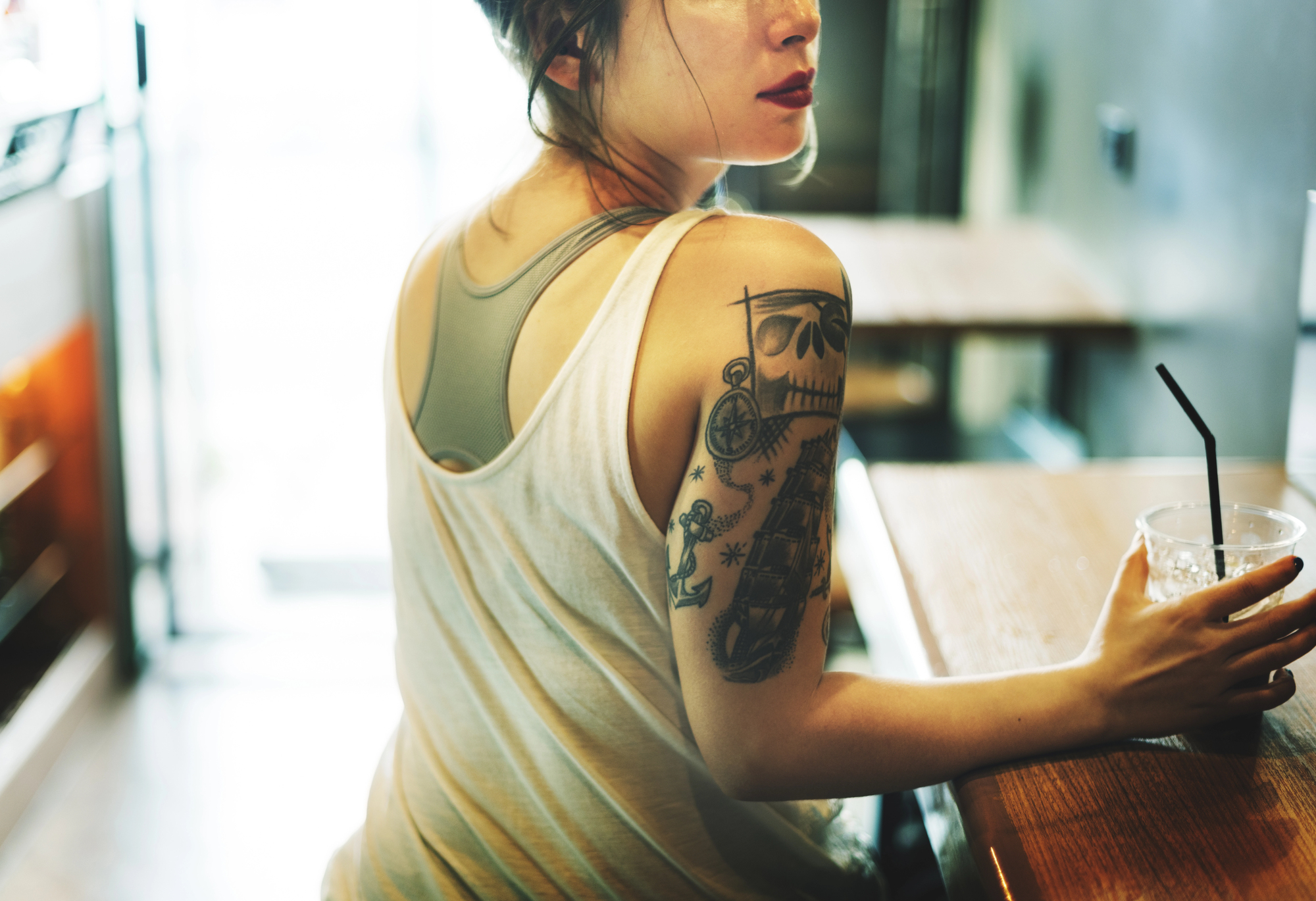 A woman with dark hair sits at a wooden table in a café, holding a drink with a straw. She is wearing a white tank top, revealing tattooed art on her left arm. The setting is warm and softly lit.