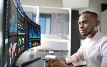 A person sitting at a desk analyzes data on a large, curved monitor displaying graphs and charts. The workspace includes a keyboard, a small plant, and a blurred window view in the background.