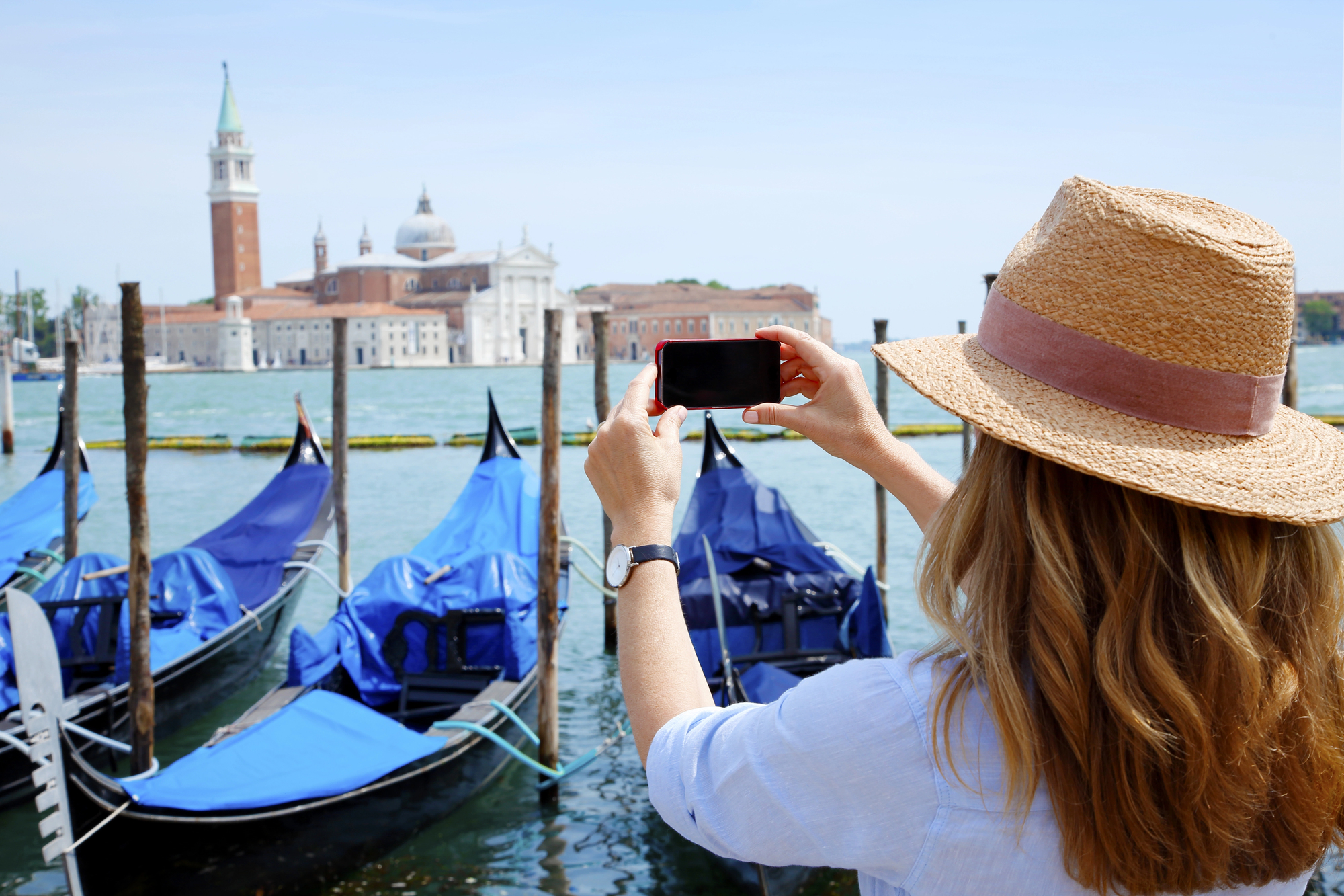 A woman wearing a straw hat is taking a photo of gondolas moored on a canal in Venice. Across the water, St. Mark's Campanile and domed buildings are visible under a clear blue sky.