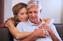 A smiling woman embraces an older man from behind as they sit on a couch. Both have their eyes closed, appearing content and relaxed. The woman has long hair, and the man has short gray hair. They are wearing casual clothing.