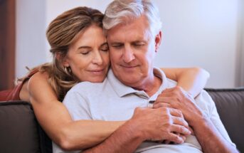 A smiling woman embraces an older man from behind as they sit on a couch. Both have their eyes closed, appearing content and relaxed. The woman has long hair, and the man has short gray hair. They are wearing casual clothing.