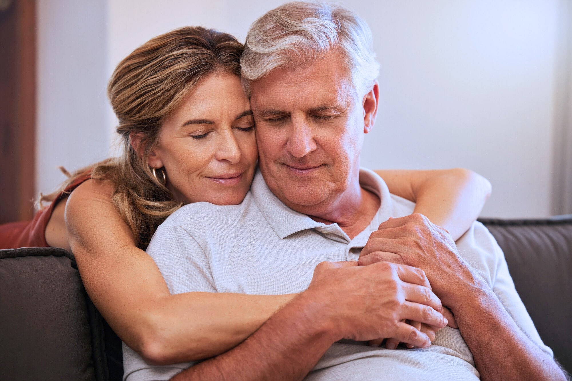 A smiling woman embraces an older man from behind as they sit on a couch. Both have their eyes closed, appearing content and relaxed. The woman has long hair, and the man has short gray hair. They are wearing casual clothing.