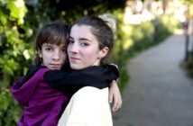 Two people embrace outdoors on a pathway surrounded by greenery. One person is a child wearing a maroon and black jacket, and the other is a young person with their hair in a bun, wearing a light-colored top. They both look towards the camera.