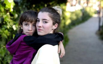 Two people embrace outdoors on a pathway surrounded by greenery. One person is a child wearing a maroon and black jacket, and the other is a young person with their hair in a bun, wearing a light-colored top. They both look towards the camera.