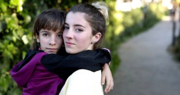 Two people embrace outdoors on a pathway surrounded by greenery. One person is a child wearing a maroon and black jacket, and the other is a young person with their hair in a bun, wearing a light-colored top. They both look towards the camera.