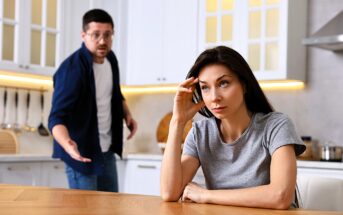 A woman sits at a kitchen table looking frustrated, resting her head on her hand. In the background, a man gestures animatedly with an open mouth, appearing to speak. The kitchen is modern with white cabinets and stainless steel appliances.