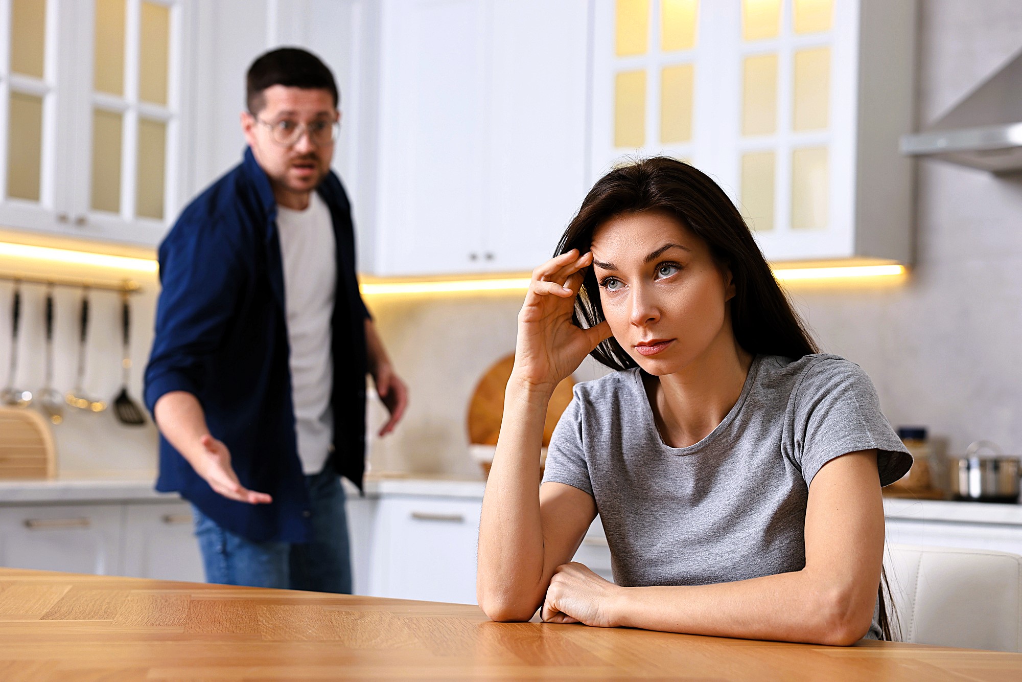 A woman sits at a kitchen table looking frustrated, resting her head on her hand. In the background, a man gestures animatedly with an open mouth, appearing to speak. The kitchen is modern with white cabinets and stainless steel appliances.