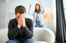 A man sits on a couch with his head in his hands, appearing stressed or upset. In the background, a woman stands with her arms raised in frustration. They are in a well-lit living room.