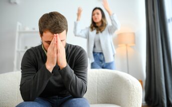 A man sits on a couch with his head in his hands, appearing stressed or upset. In the background, a woman stands with her arms raised in frustration. They are in a well-lit living room.