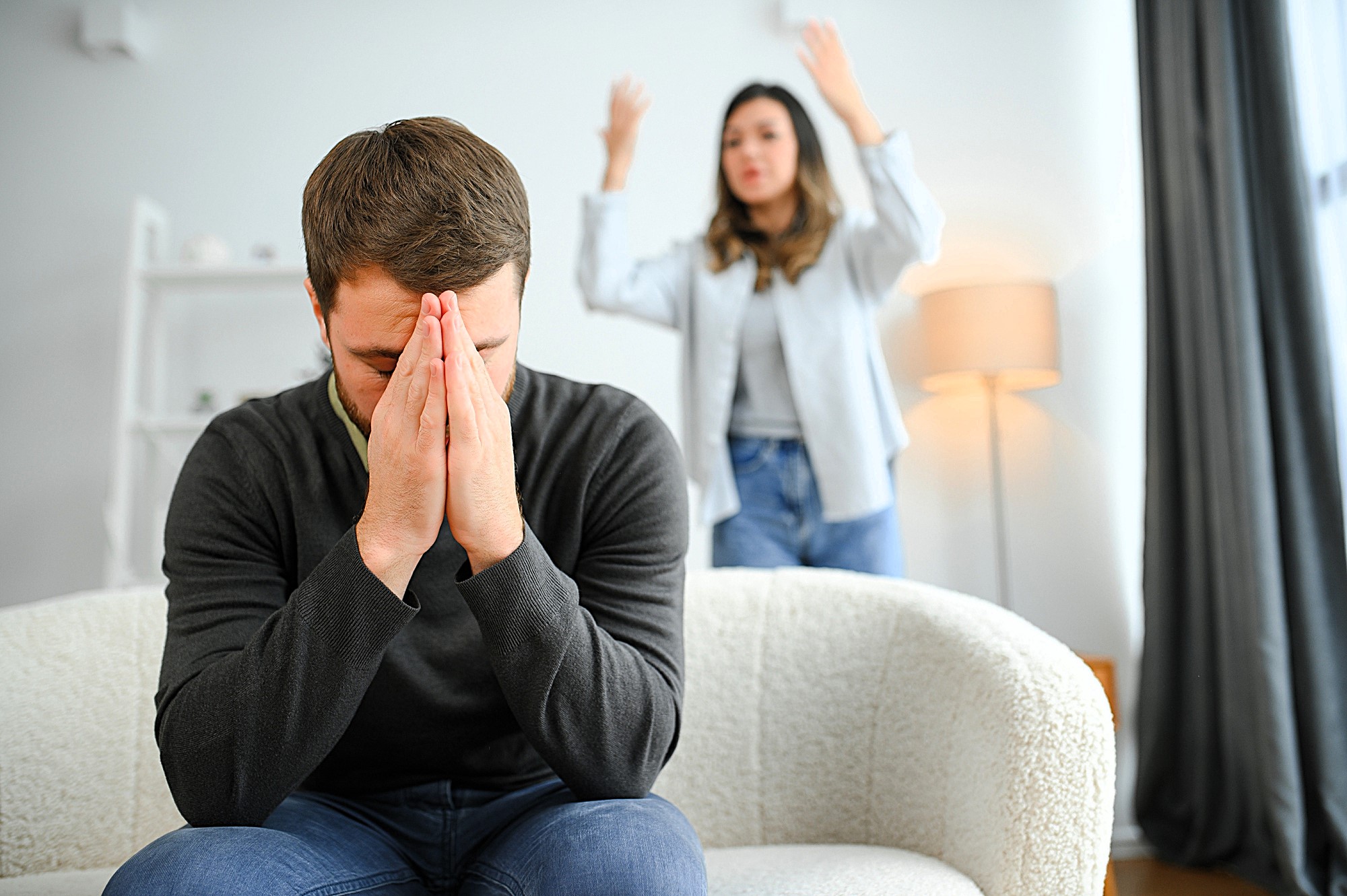 A man sits on a couch with his head in his hands, appearing stressed or upset. In the background, a woman stands with her arms raised in frustration. They are in a well-lit living room.