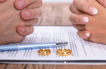 Two pairs of hands rest on a table with a document between them. A blue pen and two golden wedding rings are placed on the paper. The scene suggests a legal or personal discussion, possibly involving marriage or divorce.