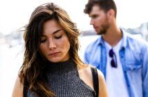 A woman in a gray sleeveless top looks down with a pensive expression. A man in a blue denim shirt and white T-shirt stands behind her out of focus, adding depth to the outdoor scene.