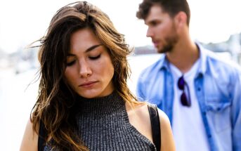 A woman in a gray sleeveless top looks down with a pensive expression. A man in a blue denim shirt and white T-shirt stands behind her out of focus, adding depth to the outdoor scene.