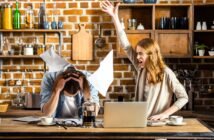 A woman angrily gestures with her arm raised while yelling at a man sitting at a kitchen table, holding his head. Papers are scattered around, and a laptop and coffee pot are on the table. The setting is a rustic kitchen with brick walls and wooden shelves.