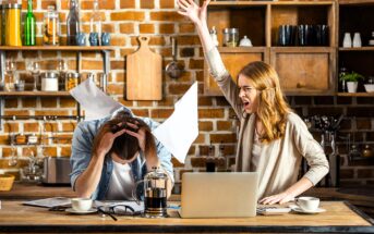 A woman angrily gestures with her arm raised while yelling at a man sitting at a kitchen table, holding his head. Papers are scattered around, and a laptop and coffee pot are on the table. The setting is a rustic kitchen with brick walls and wooden shelves.