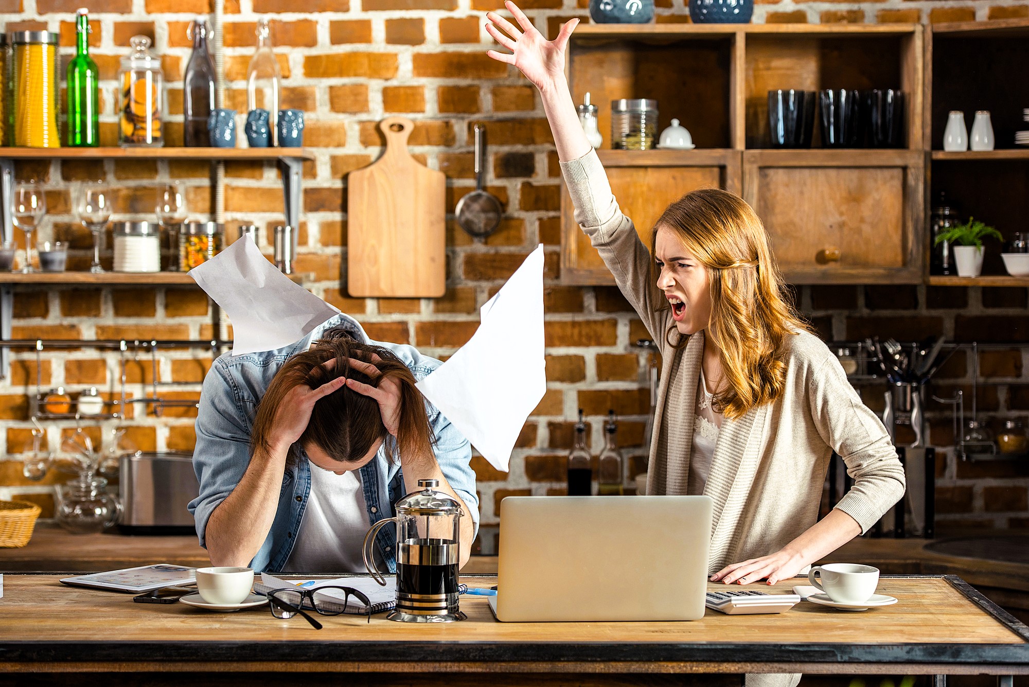 A woman angrily gestures with her arm raised while yelling at a man sitting at a kitchen table, holding his head. Papers are scattered around, and a laptop and coffee pot are on the table. The setting is a rustic kitchen with brick walls and wooden shelves.