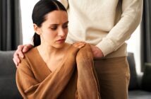 A woman in a brown sweater sits with a concerned expression as someone in a beige outfit stands behind her, gently touching her shoulder. The setting appears to be a living room.