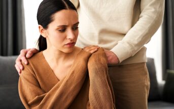 A woman in a brown sweater sits with a concerned expression as someone in a beige outfit stands behind her, gently touching her shoulder. The setting appears to be a living room.