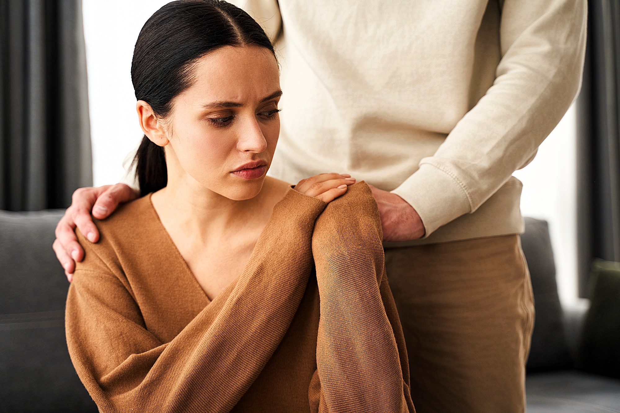 A woman in a brown sweater sits with a concerned expression as someone in a beige outfit stands behind her, gently touching her shoulder. The setting appears to be a living room.